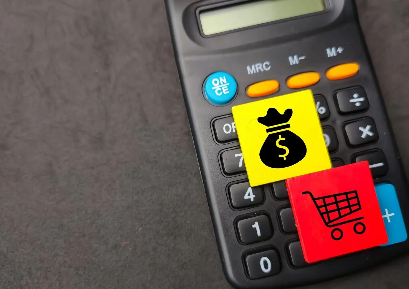 Detailní záběr dvou barevných dřevěných čtverců s ikonami nakupování a peněz na černé kalkulačce.